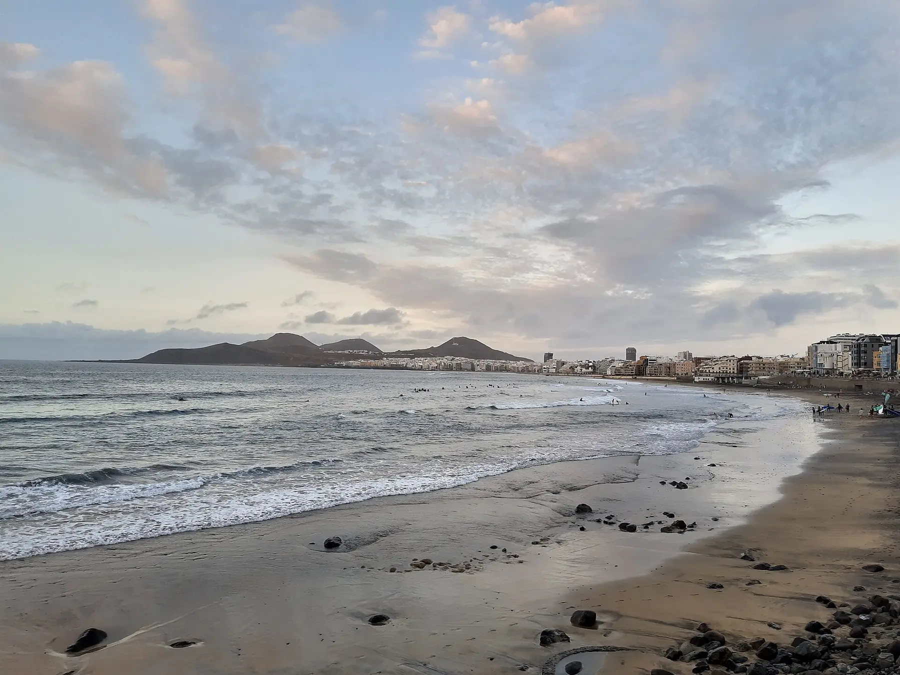 La playa de Las Canteras, en Las Palmas de Gran Canaria, con el mar, la arena oscura y la ciudad al fondo bajo un cielo de nubes.